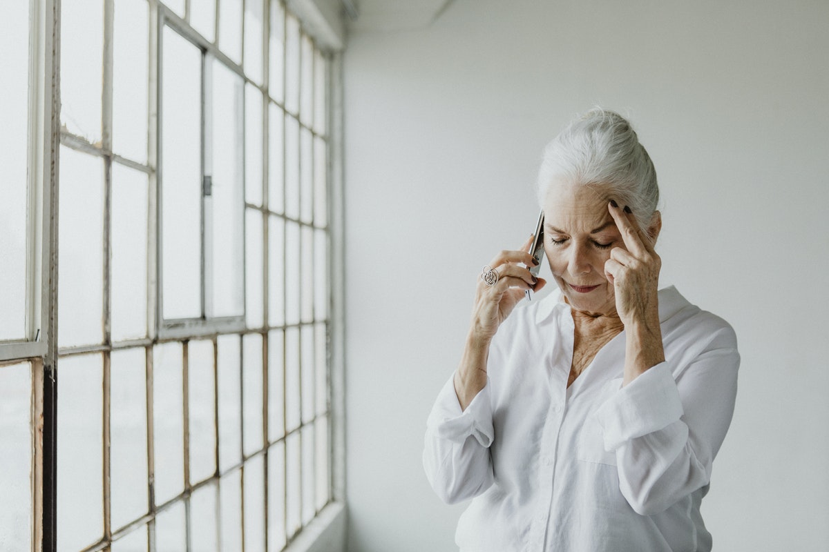 Stressed senior woman talking on a phone by the window in a white room
