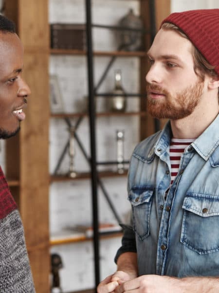 People and interracial friendship concept. Candid shot of two stylish male best friends of different races standing opposite each other and talking against modern restautant interior background
