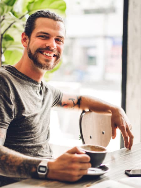 Handsome bearded hipster man relaxing while sitting on table in Young smiling man holding a cup of coffee after his long term mental health treatment in Georgetown, TX