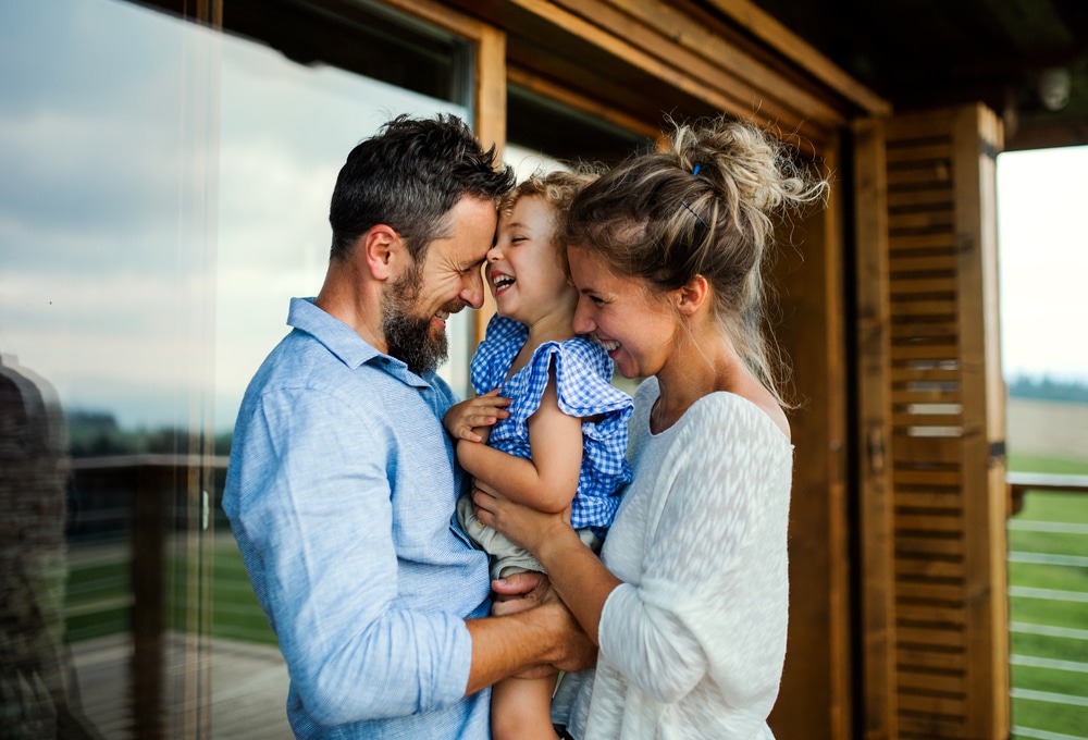 Happy family with small daughter standing on patio of wooden cabin, holiday in nature concept