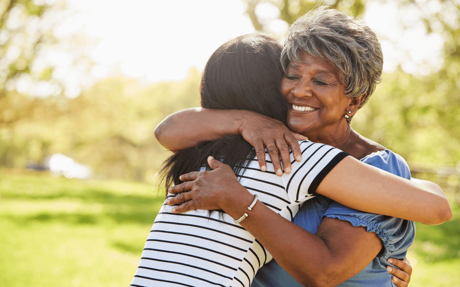 Older woman and younger woman embracing warmly outdoors in a grassy setting