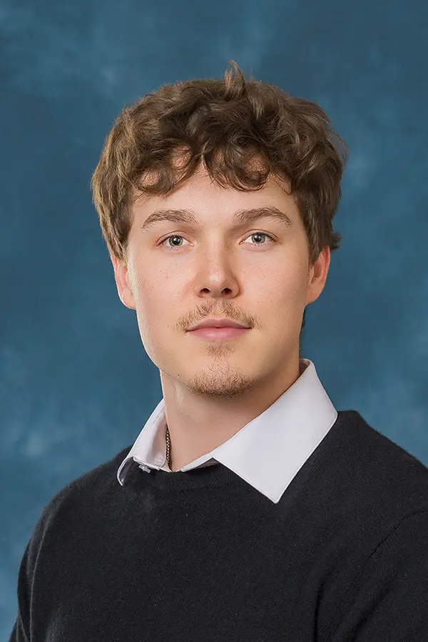 Professional headshot of a young man with curly brown hair and light facial hair, wearing a dark sweater over a light collared shirt against a blue studio background.