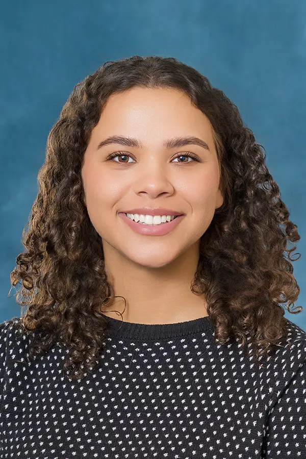 Professional headshot of a smiling woman with curly dark hair, wearing a black patterned sweater against a blue studio background.