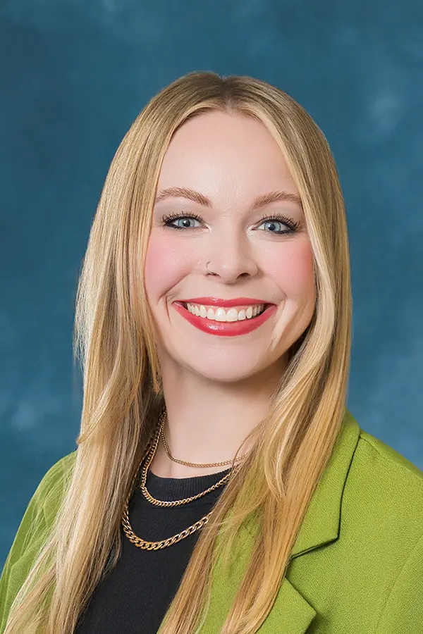 Professional headshot of a smiling woman with long blonde hair, wearing a green blazer over a black top against a blue studio background.
