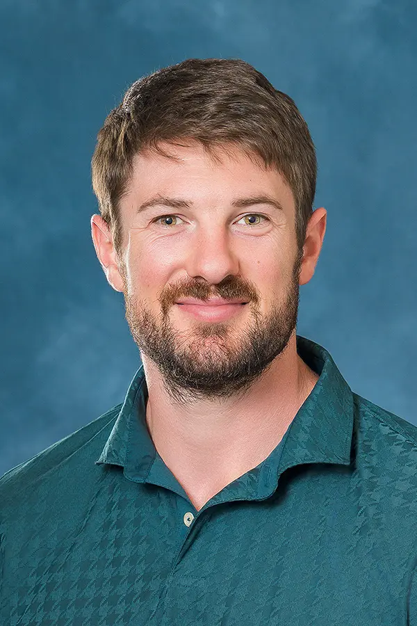 Professional headshot of a smiling man with short brown hair and a beard, wearing a teal collared shirt against a blue studio background.